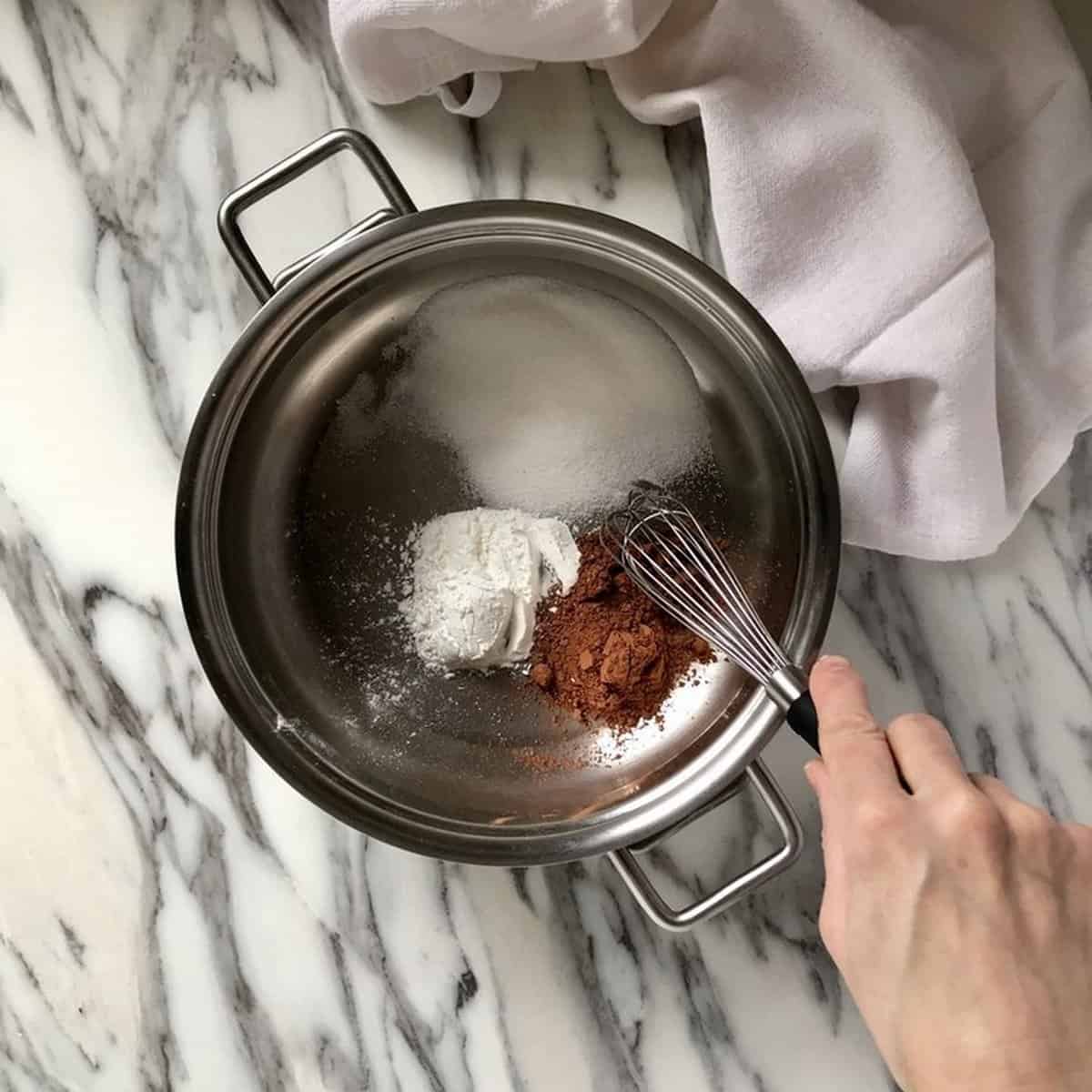 The dry ingredients to make chocolate pudding are whisked together in a saucepan. These include cornstarch, sugar and cocoa powder.
