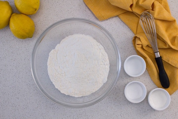The dry ingredients needed to make this lemon loaf recipe are in bowls, next to a whisk set on a yellow tea towel.