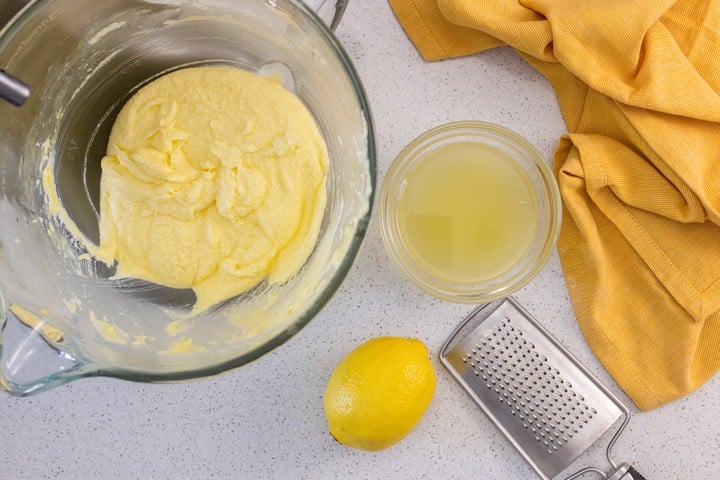An overhead shot of the lemon loaf batter in the bowl of the stand mixer next to a bowl of lemon juice, a lemon zester and a lemon.