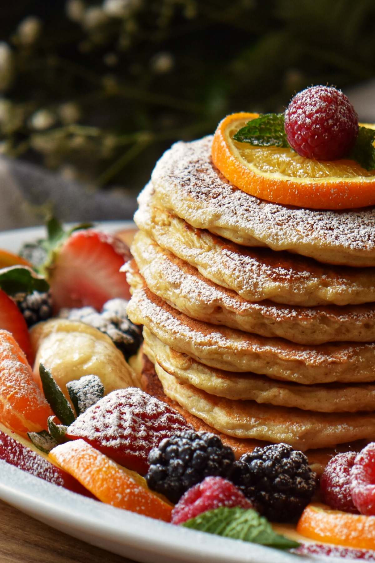 A stack of oatmeal pancakes dusted with icing sugar, garnished with a slice of orange and a raspberry.