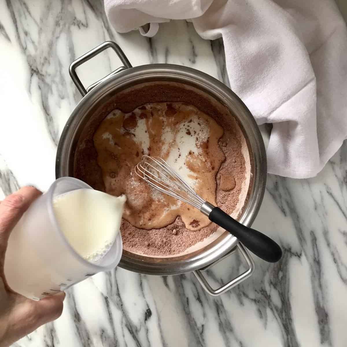 Milk is being poured in a large saucepan to be combined with the dry ingredients to make chocolate pudding.