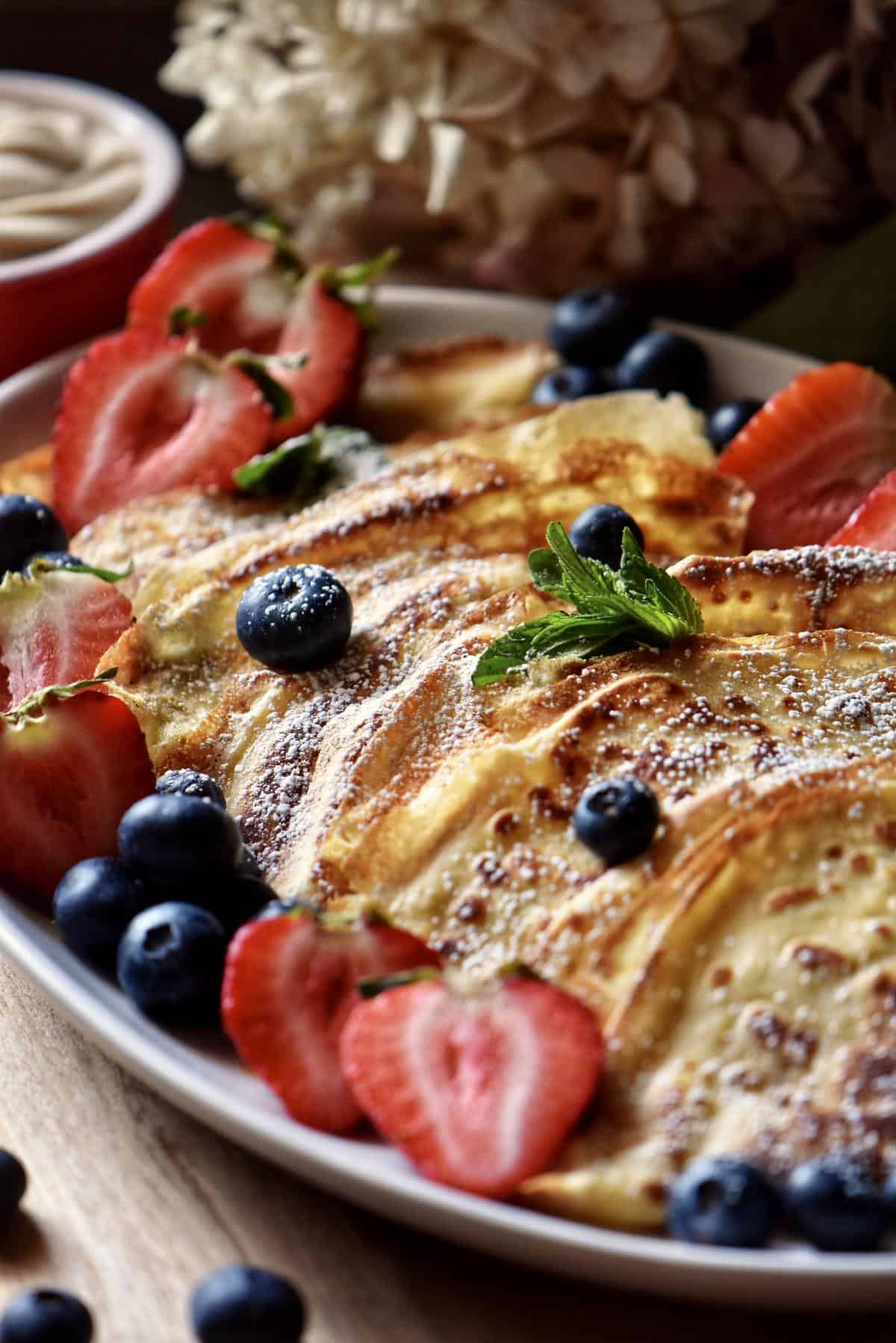 Folded crepes on a white serving platter garnished with fresh berries and icing sugar.