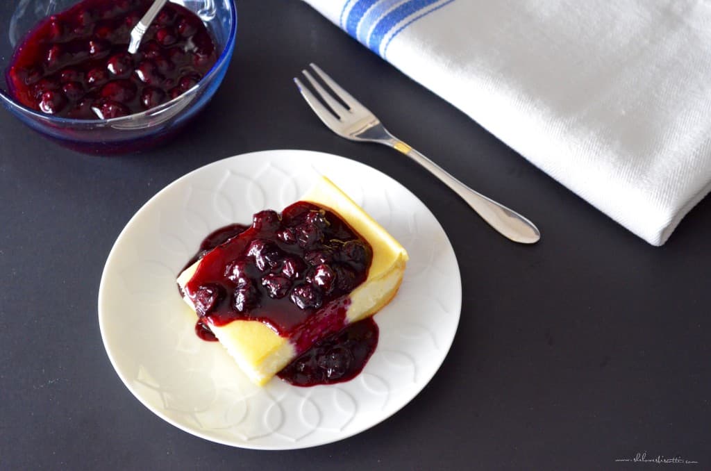 An overhead photo if a square piece of baked cheese blitzes next to a bowl of blueberry sauce.