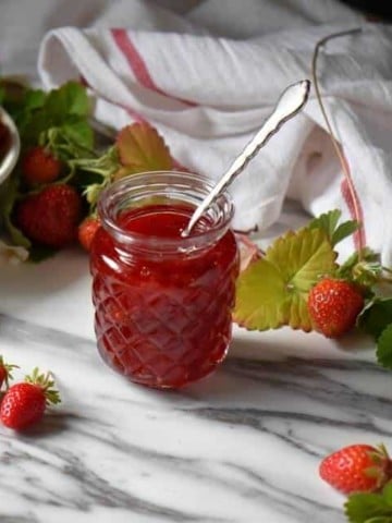 Strawberry preserves in a jar, surrounded by fresh strawberries and leaves.