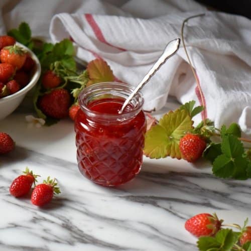 Strawberry preserves in a jar, surrounded by fresh strawberries and leaves.