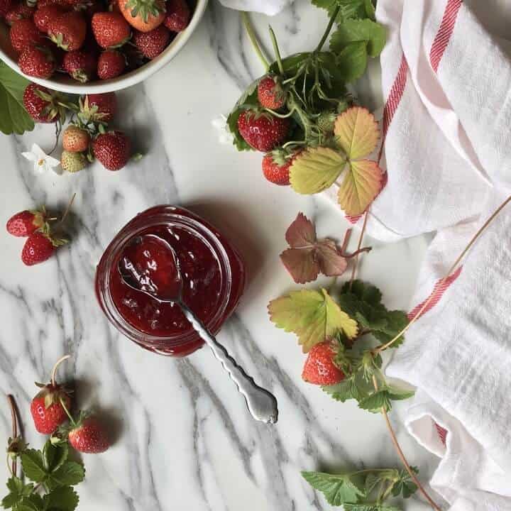 An overhead shot of a jar of strawberry jam surrounded by fresh strawberries and strawberry leaves.