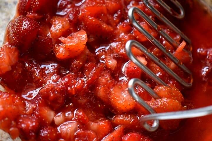 A potato masher is crushing fresh strawberries.