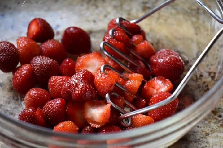 Fresh strawberries in a bowl about to be crushed by a potato masher.