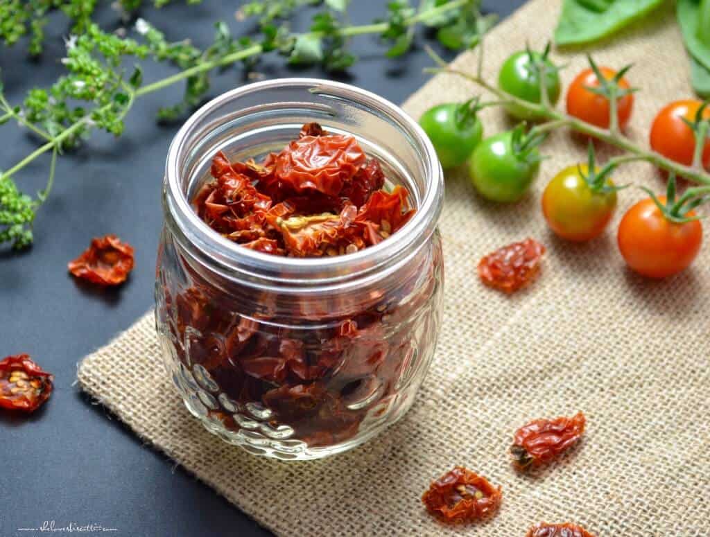 A mason jar filled with dehydrated tomatoes surrounded with fresh cherry tomatoes and oregano.