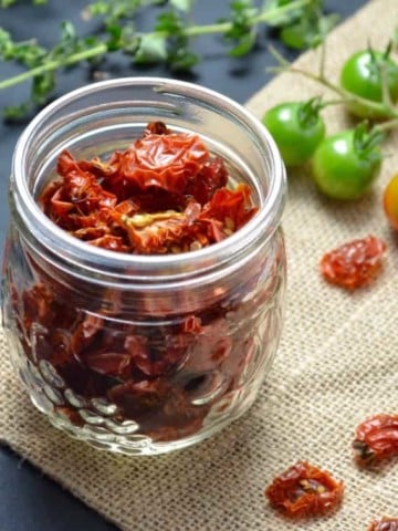 A mason jar filled with dehydrated tomatoes surrounded with fresh cherry tomatoes and oregano.