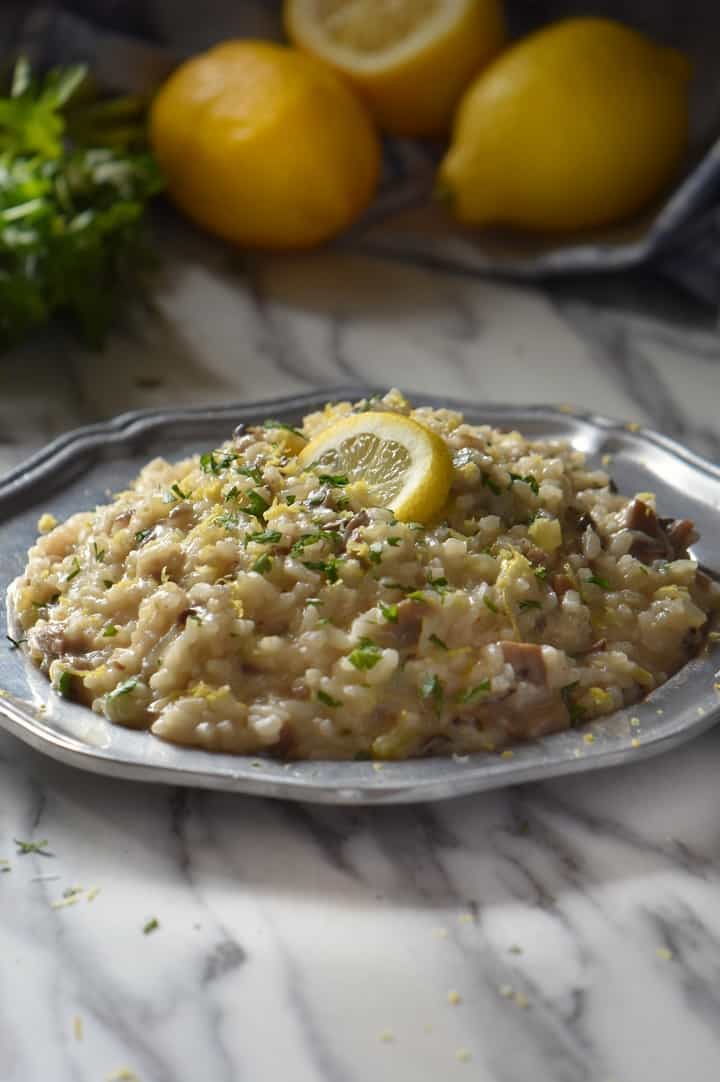 A plate of creamy mushroom risotto, topped with parsley. In the background, lemons and fresh Italian parsley can be seen.