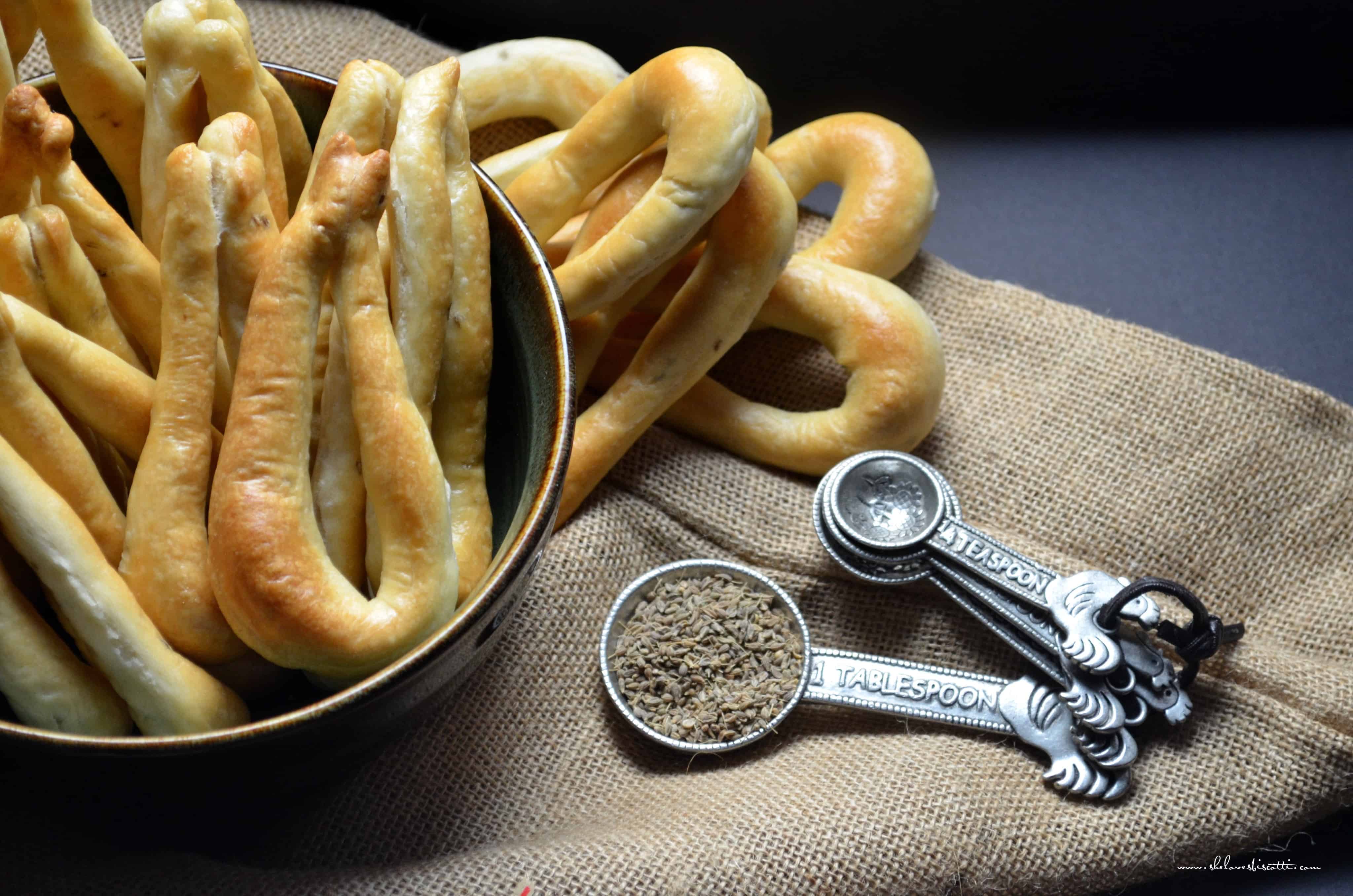 A bowl of taralli next to measuring spoons filled with anise seeds.