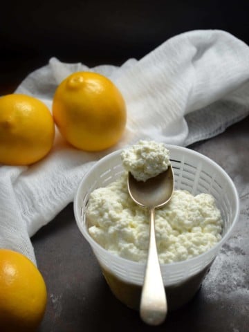 A spoonful of fresh homemade ricotta cheese placed on a cheese basket surrounded by lemons.
