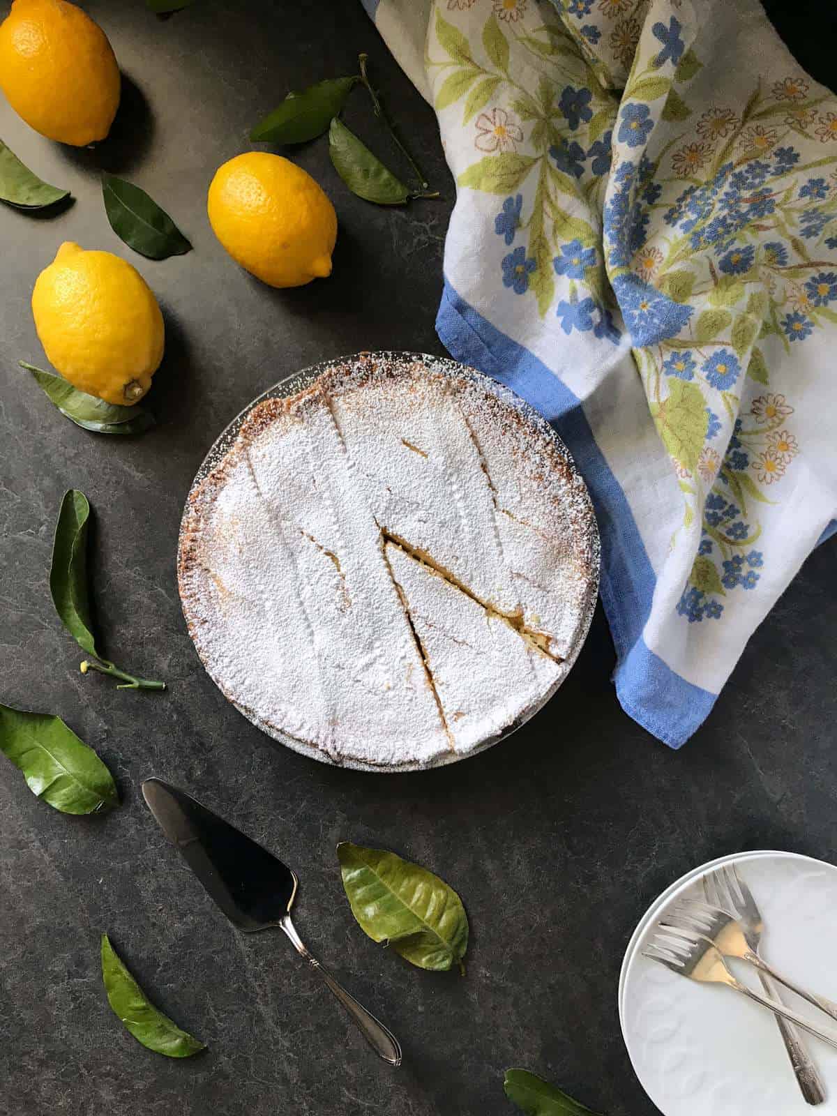 An overhead shot of a ricotta pie, dusted with icing sugar and surrounded by lemons and a floral tea towel.