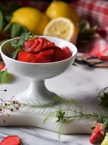 Lemons and fresh strawberries surround a white dish of macerated berries.