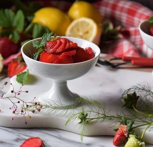 Lemons and fresh strawberries surround a white dish of macerated berries.