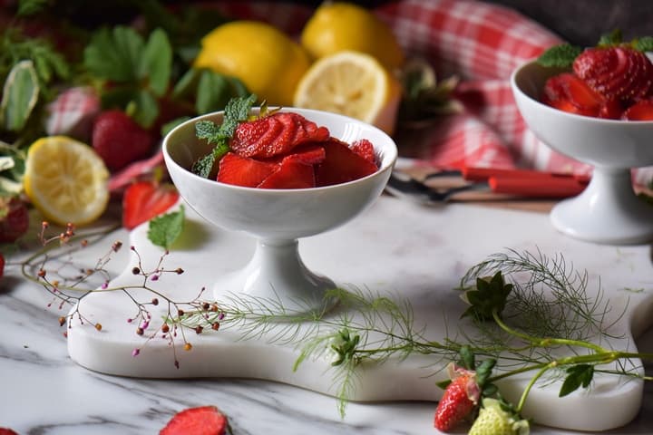 Lemons and fresh strawberries surround a white dish of macerated berries.