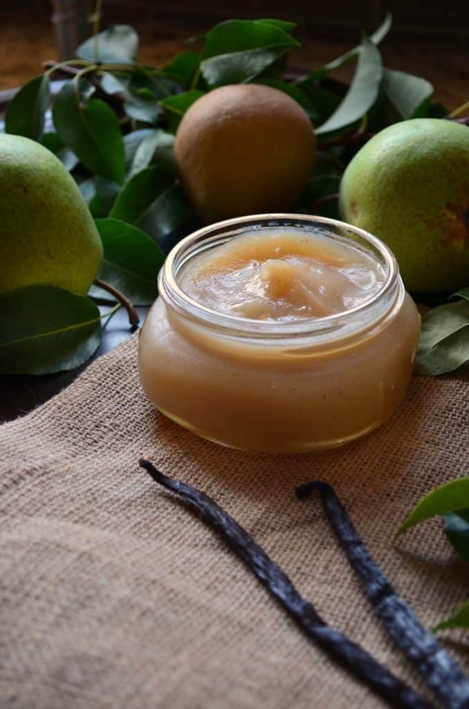 The Vanilla Pear Butter is seen in a wide open canning jar, surrounded by fresh pears.