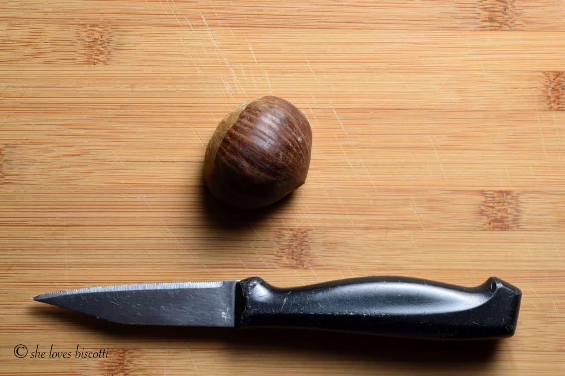 An overhead shot of a small knife with a black handle and a chestnut on a wooden cutting board.