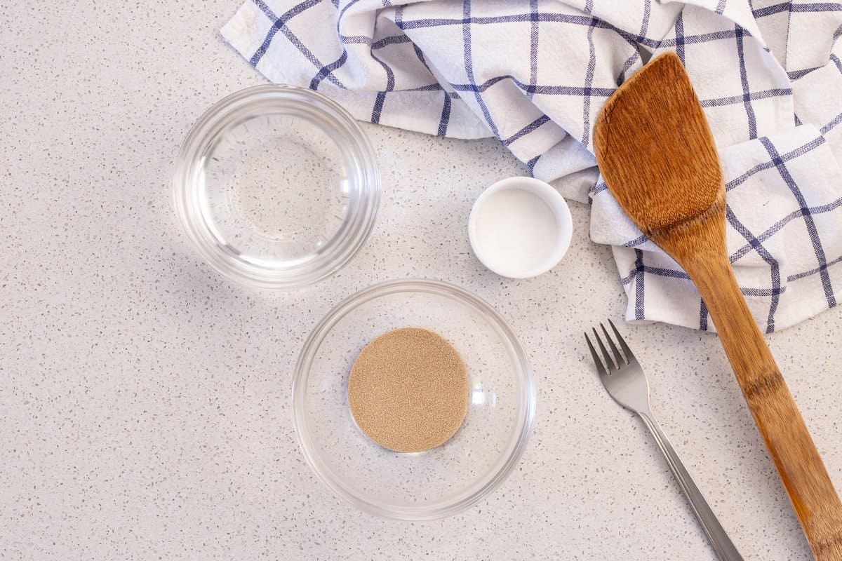 Dry yeast, salt and water in bowls, next to a for an a large wooden spoon.