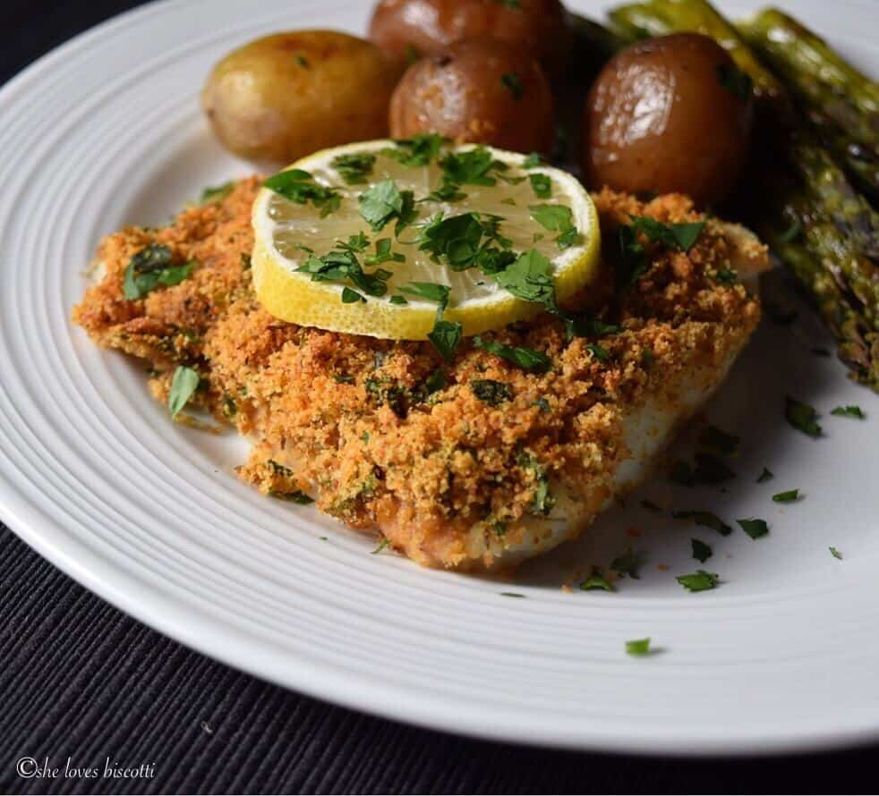 A piece of baked cod fish on a white plate topped with a slice of lemon.