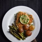 An overhead shot of baked cod fish with a side of potatoes and asparagus on a white plate.