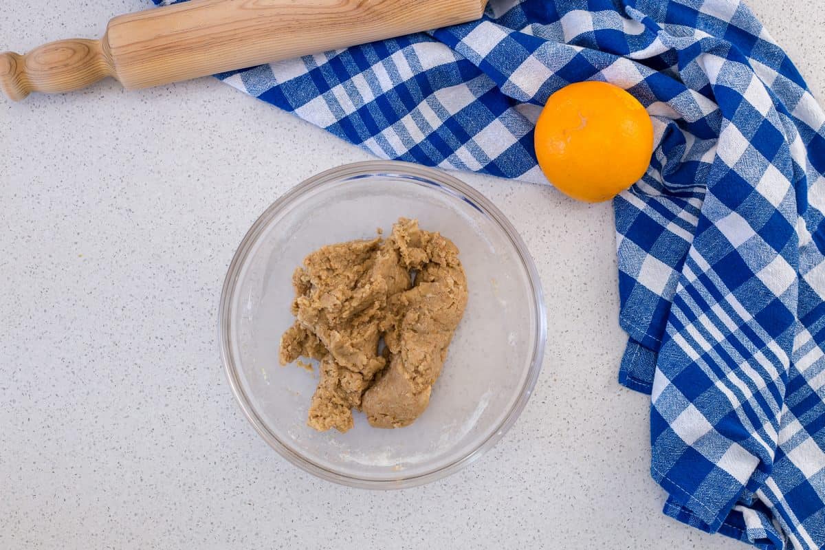 A ball of dough placed at the bottom of a bowl. Also pictured are a checkered blue tea towel, a rolling pin and a lemon.