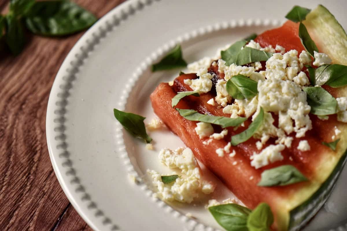 An overhead shot of a grilled watermelon topped with cheese and fresh basil leaves.