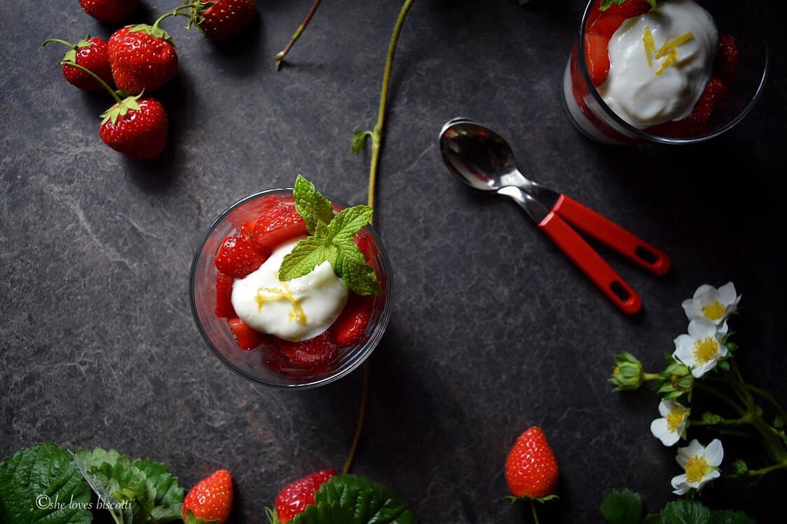 An overhead shot of a strawberry parfait topped with mint. Also visible are strawberry blooms and freshly picked strawberries with their stems.