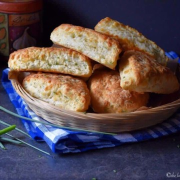 Fluffy garlic scape cheddar biscuits in a wicker basket, set on a blue checkered tea towel.