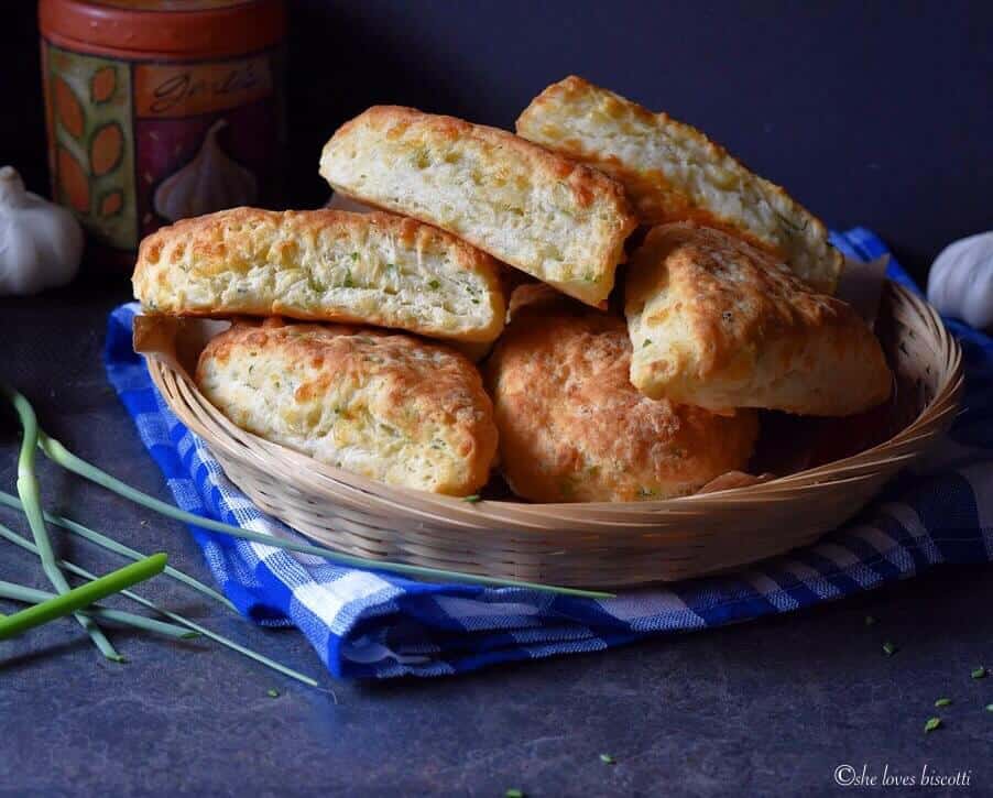 Fluffy garlic scape cheddar biscuits in a wicker basket, set on a blue checkered tea towel.