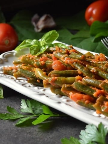Green Beans with Tomatoes on a white ceramic dish surrounded by fresh tomatoes and garlic.