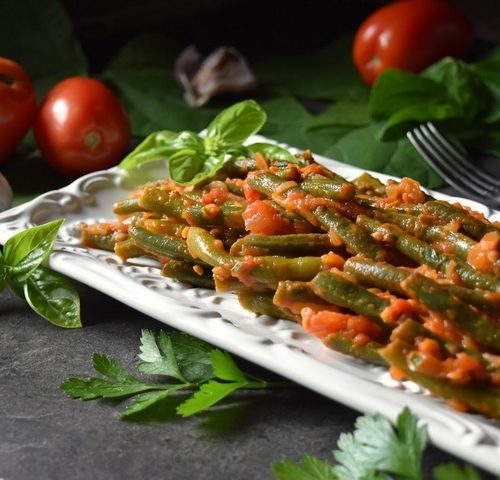 Green Beans with Tomatoes on a white ceramic dish surrounded by fresh tomatoes and garlic.
