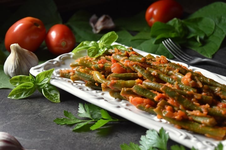 Green Beans with Tomatoes on a white ceramic dish surrounded by fresh tomatoes and garlic.