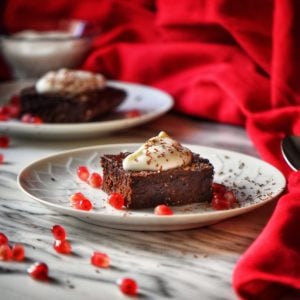 A close up shot of a square piece of Healthy Chocolate Brownie topped with whipped ricotta and surrounded with pomegranate arils.