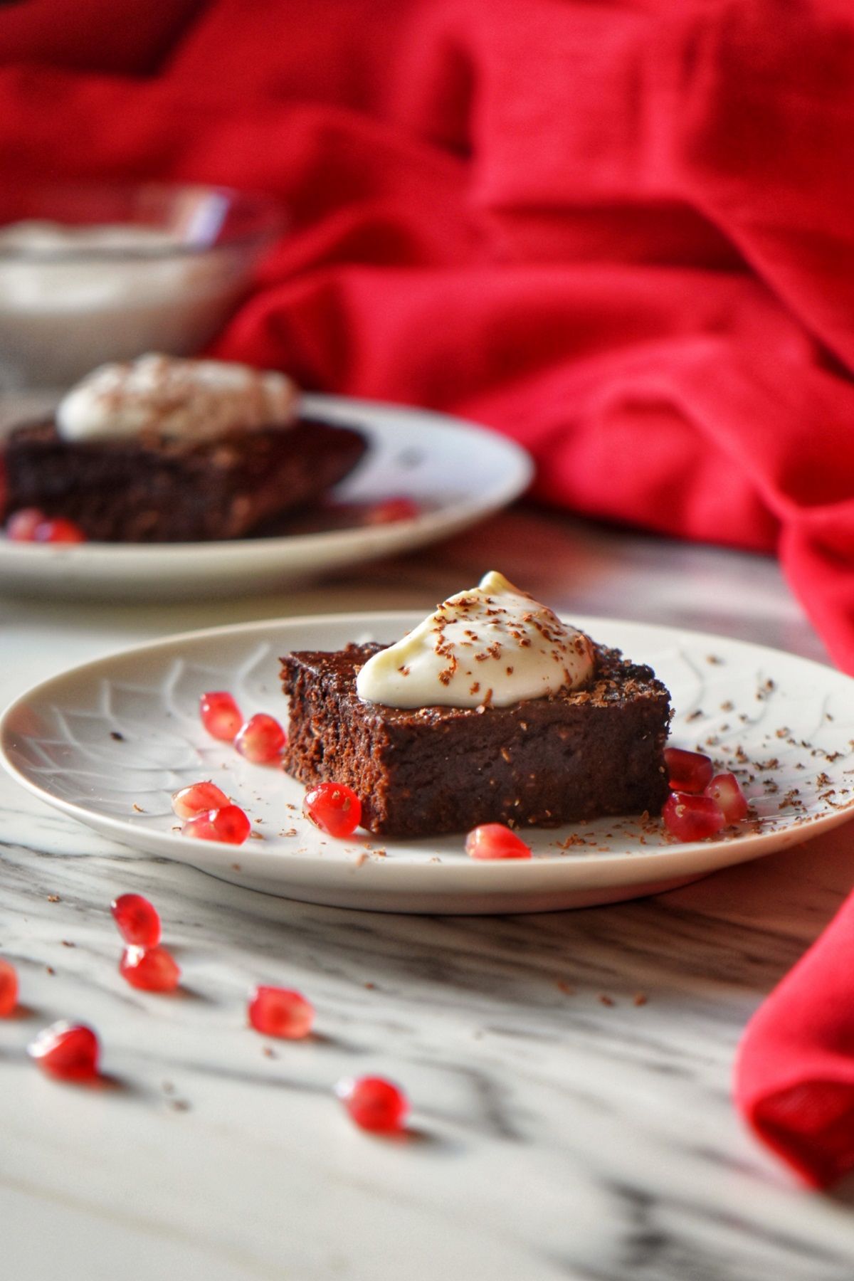 A piece of healthy chocolate brownie on a white plate surrounded by pomegranate arils.