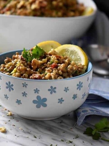 A close up a small flowery blue bowl piled high with lentil salad. Walnuts, parsley, fresh chives and a lemon wedge surrounds the bowl.