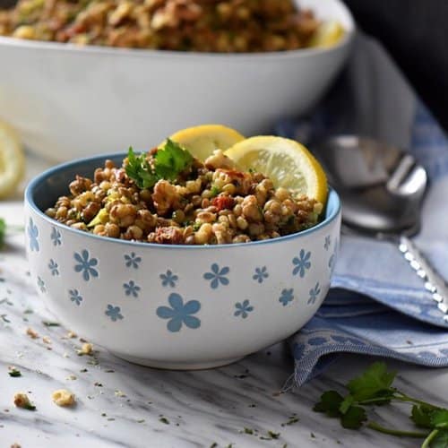 A close up a small flowery blue bowl piled high with lentil salad. Walnuts, parsley, fresh chives and a lemon wedge surrounds the bowl.