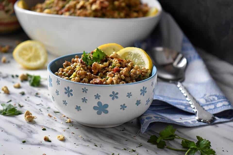 A close up a small flowery blue bowl piled high with lentil salad. Walnuts, parsley, fresh chives and a lemon wedge surrounds the bowl.