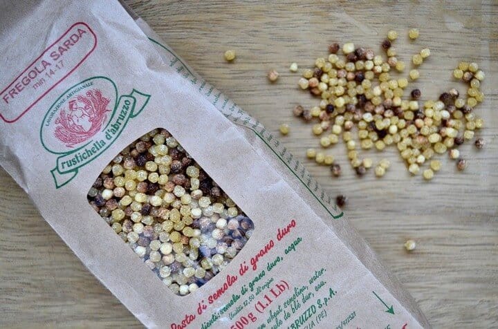 An overhead shot of the packaged fregola along with a handful of fregola on a wooden board.