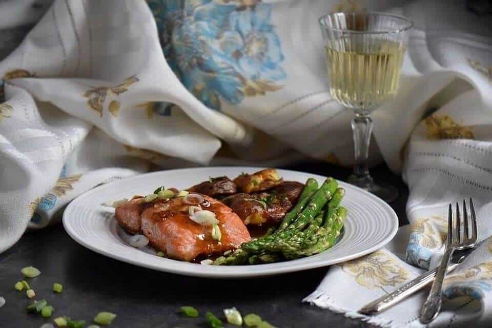 A piece of glazed salmon served on a white plate along with sauteed asparagus and smashed potatoes.