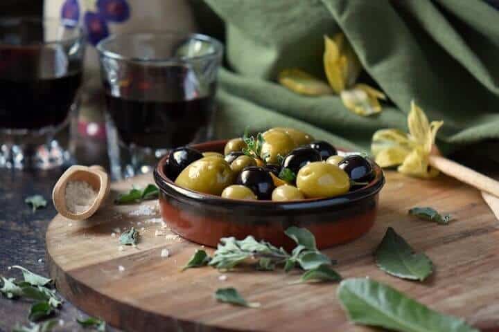 Marinated olives with a glass of wine in the background, surrounded by a few fresh oregano leaves. 