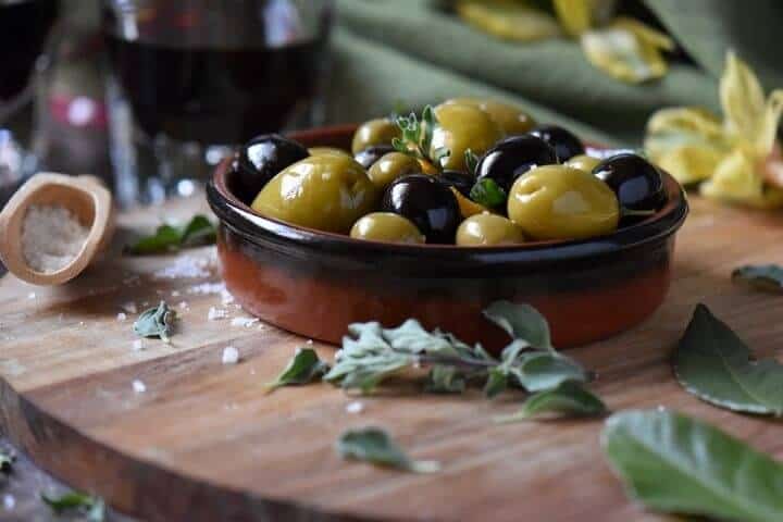 Marinated olives, of the green and black variety in a small dish, surrounded by oregano and salt.