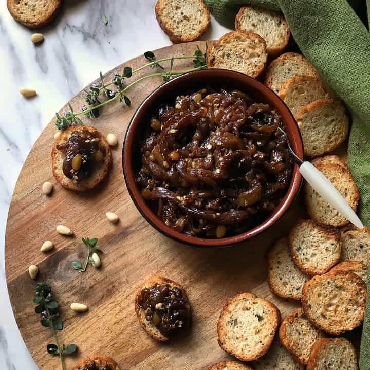 An overhead shot of eggplant caponata in a round dish, surrounded by sliced crispy baguette and scattered pine nuts.