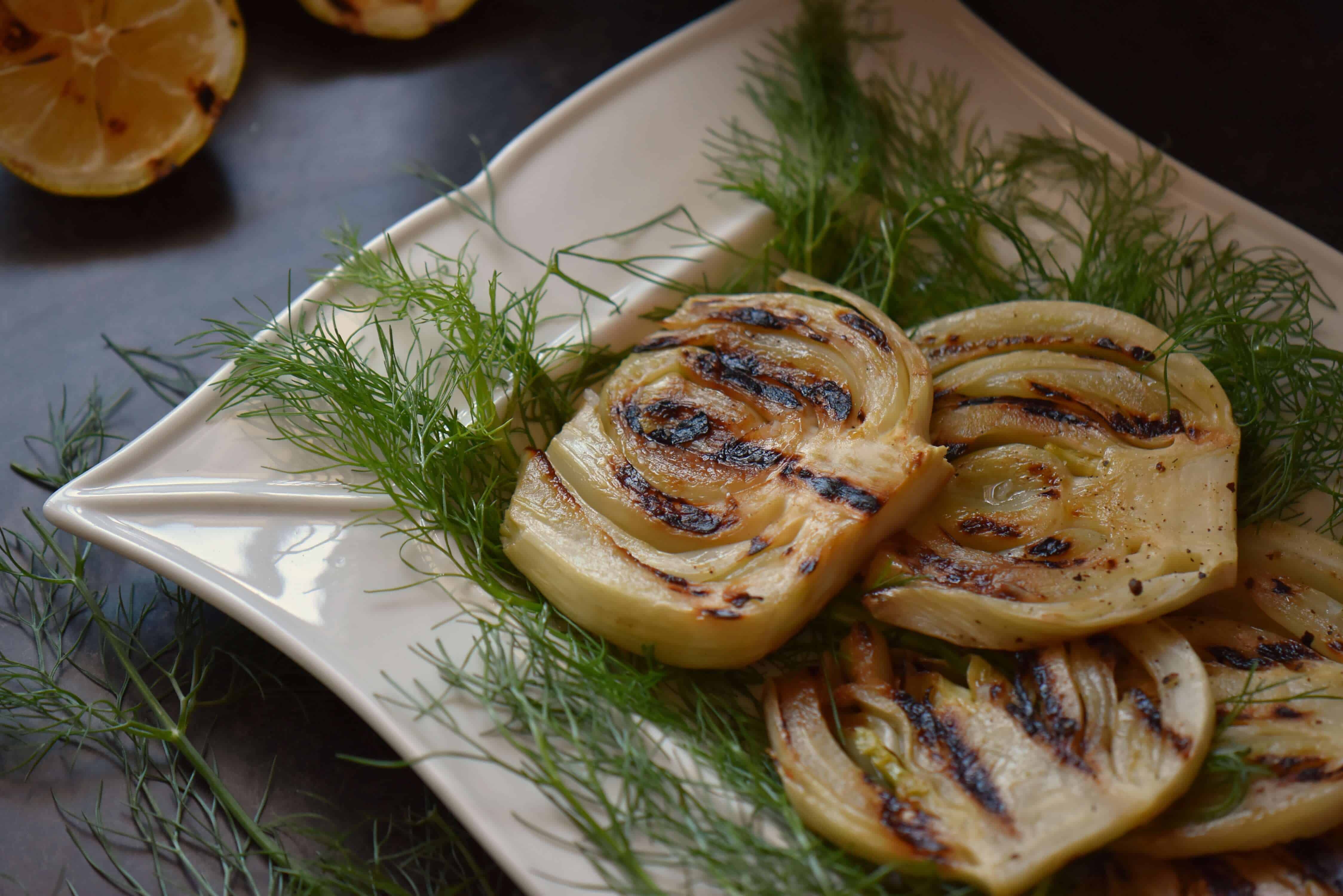 A few slices of grilled fennel, placed on some fennel fronds on a white serving dish.