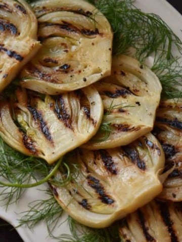 Grilled fennel placed on fennel fronds on a white serving platter.