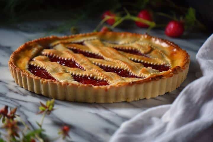 The Italian Jam Tart on a white marbled surface, surrounded by fresh strawberries.