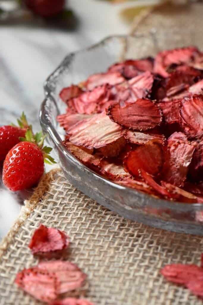 Oven dried strawberries, in a round glass dish, surrounded by a few fresh strawberries.