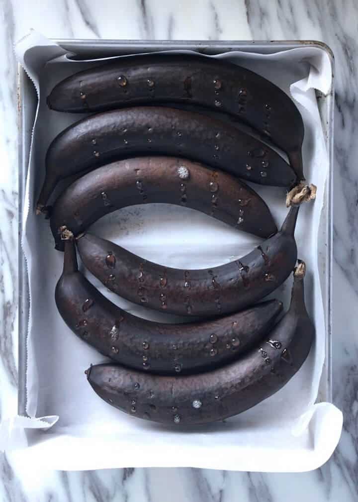 Six black skinned bananas on a parchment lined baking tray.