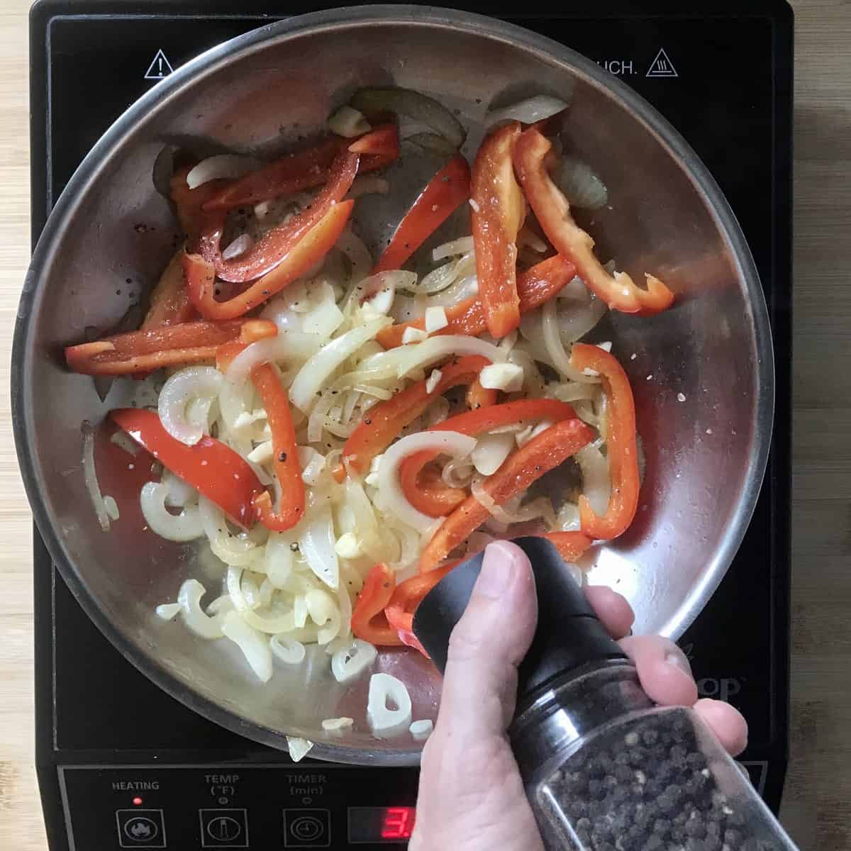 Sliced onions and red peppers are being sauteed in a skillet and seasoned with a grinding of black pepper. 